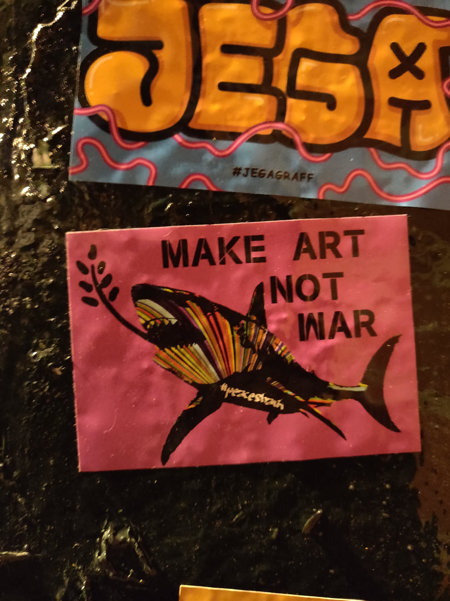 Pink square sticker featuring a stylized shark holding an olive branch in its mouth. The text "MAKE ART NOT WAR" is prominently displayed. The shark is depicted in colorful, striped patterns.