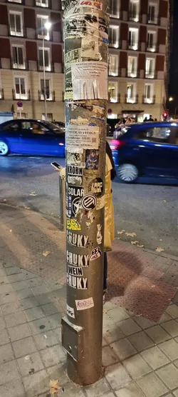 The central pole is a dense collage of various stickers and pasted papers, showcasing a vibrant mix of urban communication. The upper section is dominated by torn paper flyers, some with typed advertisements for cleaning services and private tutoring in mathematics and physics, featuring contact phone numbers. Below these, a diverse collection of graphic stickers emerges. One prominent sticker depicts an anime-style character, possibly a magical girl or witch, rendered in blues, purples, and oranges. Several bold, black-and-white stickers feature brand-like names such as 'KROHN' and 'SOLAR' in strong typography. Other graphic elements include abstract symbols, a star design with 'HSU', and a yellow graffiti-style tag. The lower part of the pole is characterized by a series of large, impactful black-and-white stickers repeating the word 'CHUKY' with the phrase 'LA LUCHA SIGUE' (The fight continues) below it, interspersed with 'WEIN'. Smaller, whimsical stickers, like a cartoonish pink frog-like creature and a stylized hand, are scattered throughout. The overall appearance is a textured, multi-layered surface reflecting the dynamic and often chaotic visual landscape of a city street.