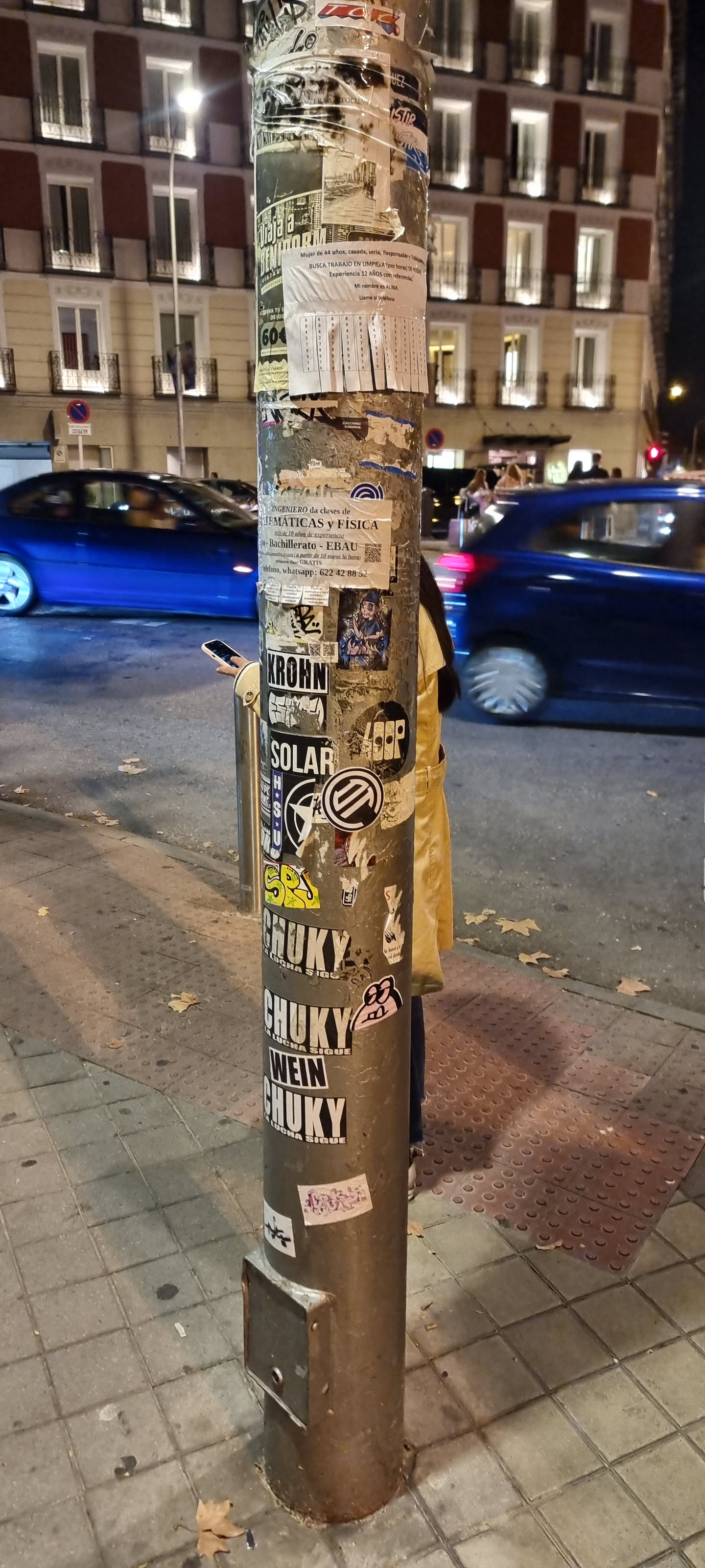 The central pole is a dense collage of various stickers and pasted papers, showcasing a vibrant mix of urban communication. The upper section is dominated by torn paper flyers, some with typed advertisements for cleaning services and private tutoring in mathematics and physics, featuring contact phone numbers. Below these, a diverse collection of graphic stickers emerges. One prominent sticker depicts an anime-style character, possibly a magical girl or witch, rendered in blues, purples, and oranges. Several bold, black-and-white stickers feature brand-like names such as 'KROHN' and 'SOLAR' in strong typography. Other graphic elements include abstract symbols, a star design with 'HSU', and a yellow graffiti-style tag. The lower part of the pole is characterized by a series of large, impactful black-and-white stickers repeating the word 'CHUKY' with the phrase 'LA LUCHA SIGUE' (The fight continues) below it, interspersed with 'WEIN'. Smaller, whimsical stickers, like a cartoonish pink frog-like creature and a stylized hand, are scattered throughout. The overall appearance is a textured, multi-layered surface reflecting the dynamic and often chaotic visual landscape of a city street.