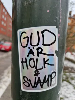 A sticker with holographic foil background. Black hand-written text says: "Gud är holk & svamp" which translates to "God is a mushroom and a hollow".