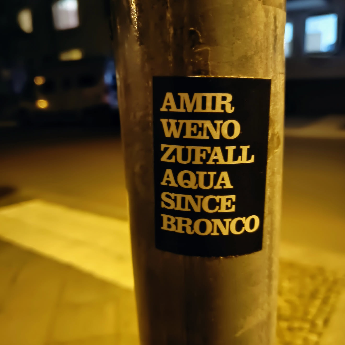 A black sticker on a metal pole with white text reading: AMIR WENO ZUFALL AQUA SINCE BRONCO. The words are stacked vertically.