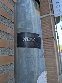 A rectangular sticker with a solid black background featuring the word 'OTYDLIG' printed in white capital letters using a bold, sans-serif font. The sticker is placed horizontally on a weathered, round metal post. The right side of the sticker shows minor wear and a small tear along its edge.