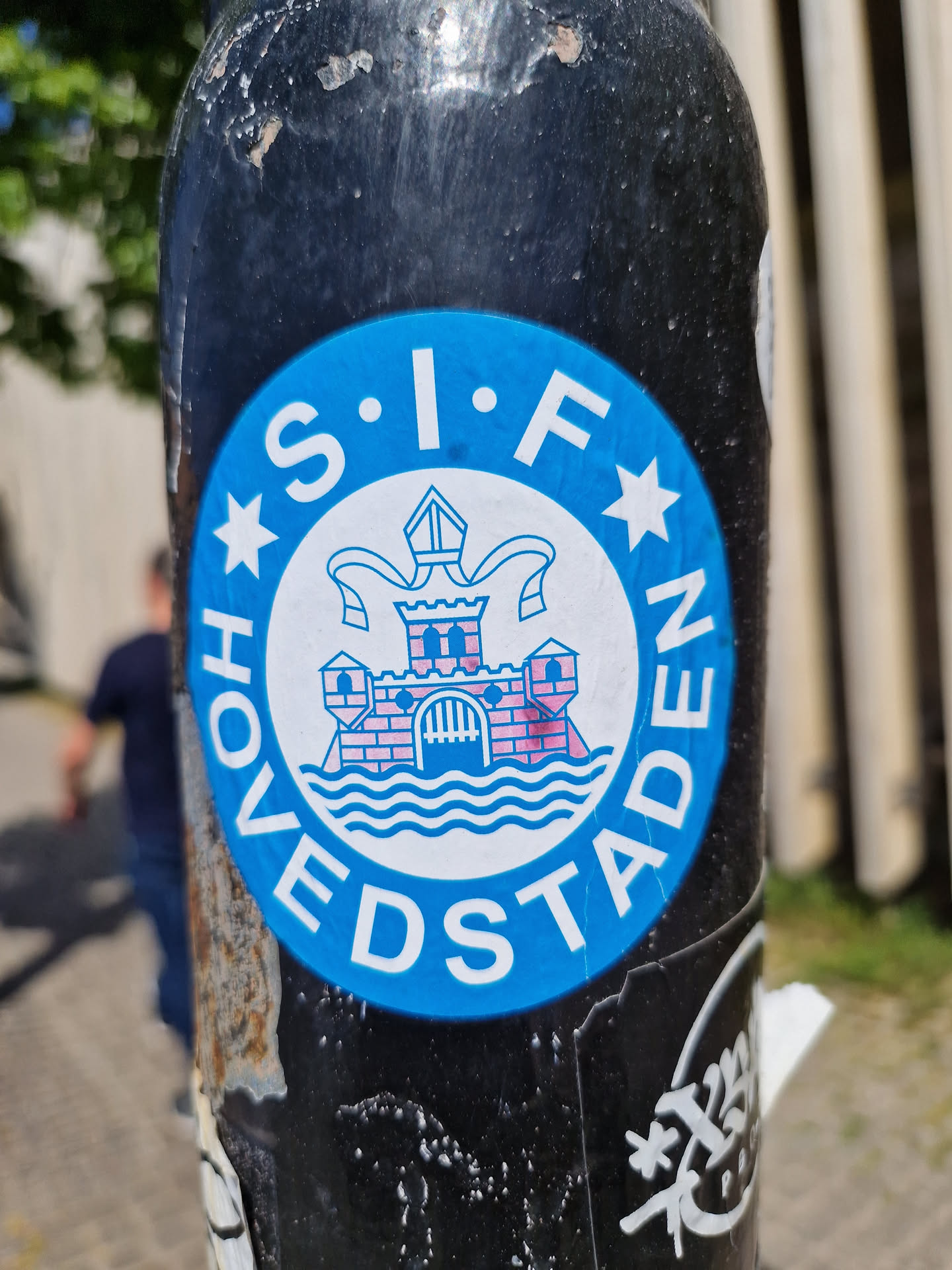 A blue circular sticker with white and pink details featuring the logo of what appears to be a Danish football club, SIF HOVEDSTADEN. The logo includes a stylized castle and wavy lines suggesting water. The sticker is affixed to a dark-colored pole, showing some wear and tear.