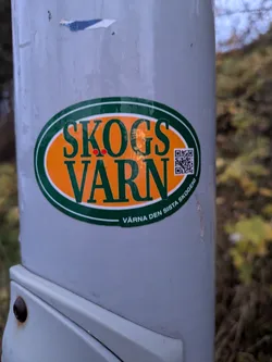 A SkogsVarn sticker affixed to a grey pole. The sticker is oval-shaped with an orange background and green border. The text 'SKOGS VARN' is displayed prominently in dark green lettering. Below this, in smaller text, is 'VARNA DEN SISTA SKOGEN' (Protect the last forest). A QR code is situated to the right of the main text. The sticker promotes environmental protection and forest conservation.
