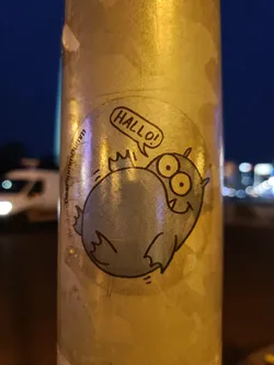 Round sticker featuring a cartoon monster with big eyes and a speech bubble saying "Hallo!" in German. The monster is light blue and has small bat-like wings. The sticker appears to be affixed to a lamppost or similar outdoor surface.