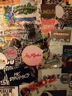 Pink round sticker with the text "Joy Ryder" in a stylized font. Part of a larger collection of stickers on a wall.