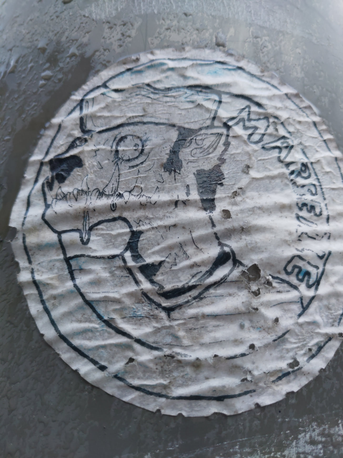 A weathered, circular sticker with a monochrome illustration of a creature's head. The style is reminiscent of dark fantasy or gothic art. The sticker shows significant signs of wear and tear, including wrinkles, creases, and some loss of material. Part of the text on the sticker is illegible due to the damage.