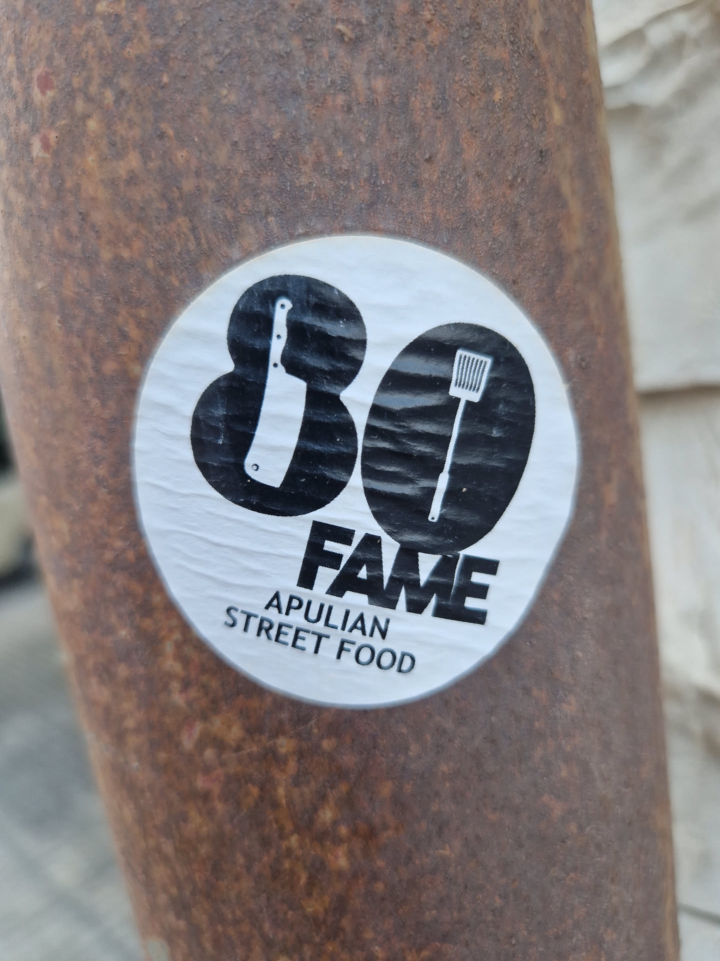A round sticker with the number 80 in the center. On each side of the number there is an icon: a meat cleaver and a spatula. Underneath the number, it says FAME in large letters and APULIAN STREET FOOD in smaller letters.