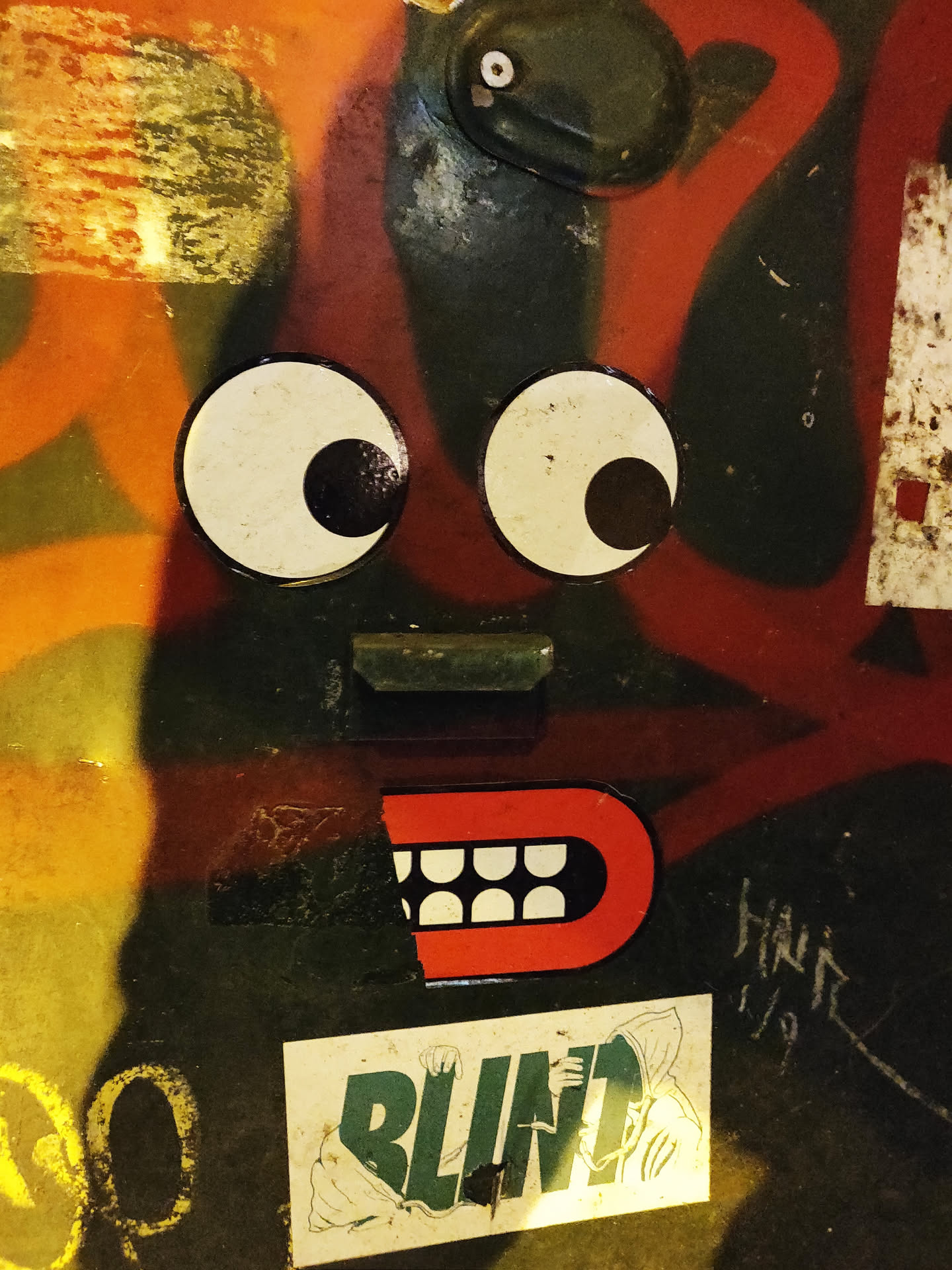 A sticker featuring a pair of large googly eyes and a red mouth with white teeth. It is adhered to a surface with various other stickers and graffiti.