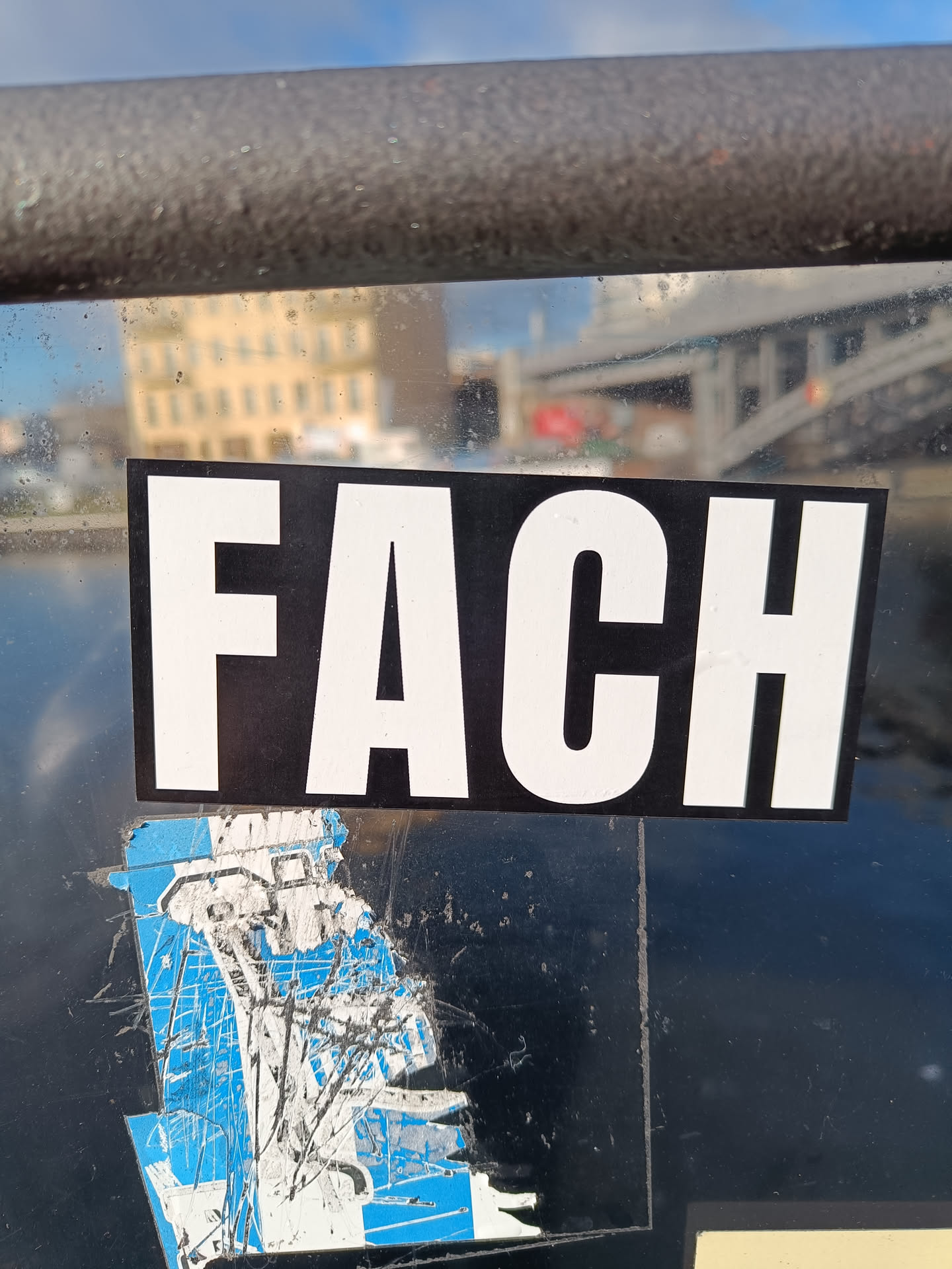 A black and white sticker with the German word "FACH" (meaning "subject", "field", or "compartment") is affixed to a transparent surface.  Part of another sticker, blue and white, is visible underneath, suggesting it may have been placed overtop. The background shows a blurred-out city scene with water.