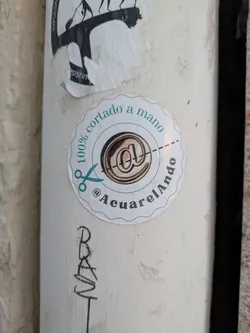 The sticker is round with a scalloped edge. The text "100% cortado a mano" (100% hand cut) runs along the top curve, and the Instagram handle @AcuarelAndo runs along the bottom. In the center is an image that looks like a stylized "@" symbol. A pair of scissors is placed as if to cut the sticker along a dotted line.