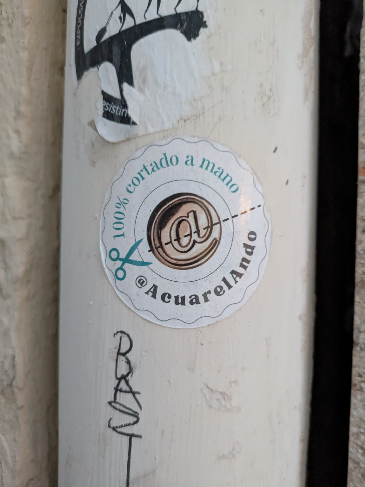 The sticker is round with a scalloped edge. The text "100% cortado a mano" (100% hand cut) runs along the top curve, and the Instagram handle @AcuarelAndo runs along the bottom. In the center is an image that looks like a stylized "@" symbol. A pair of scissors is placed as if to cut the sticker along a dotted line.