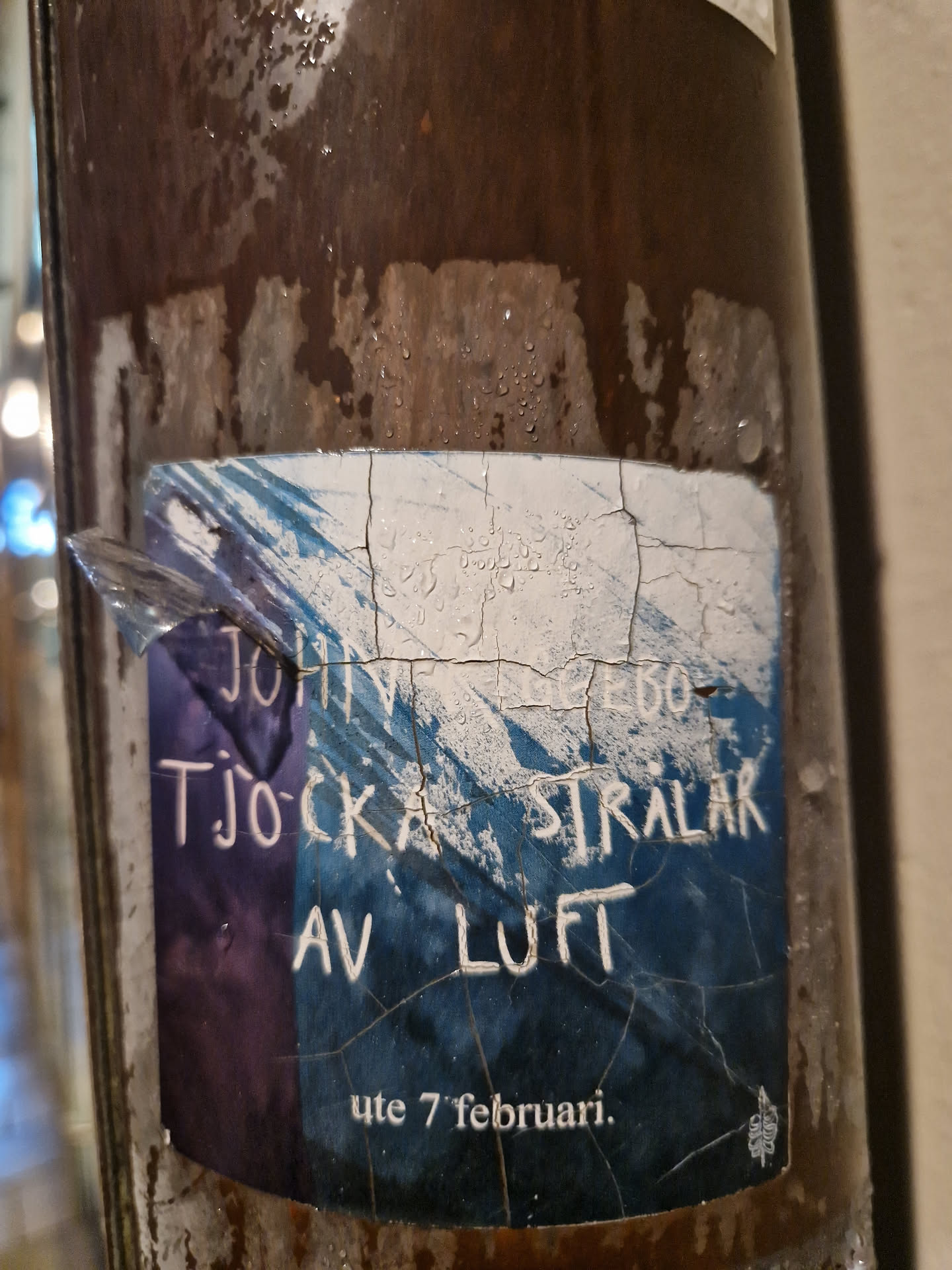 A worn-out sticker announcing an event: John Ljebø - Tjocka strålar av luft.  The date is visible: ute 7 februari (7th of February). The sticker shows some damage and weathering.