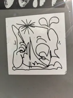 A white square sticker with a black and white line drawing of a cartoonish figure. The figure appears to be sitting or lounging, with a somewhat whimsical and abstract style. There's a small starburst element near the figure's head.