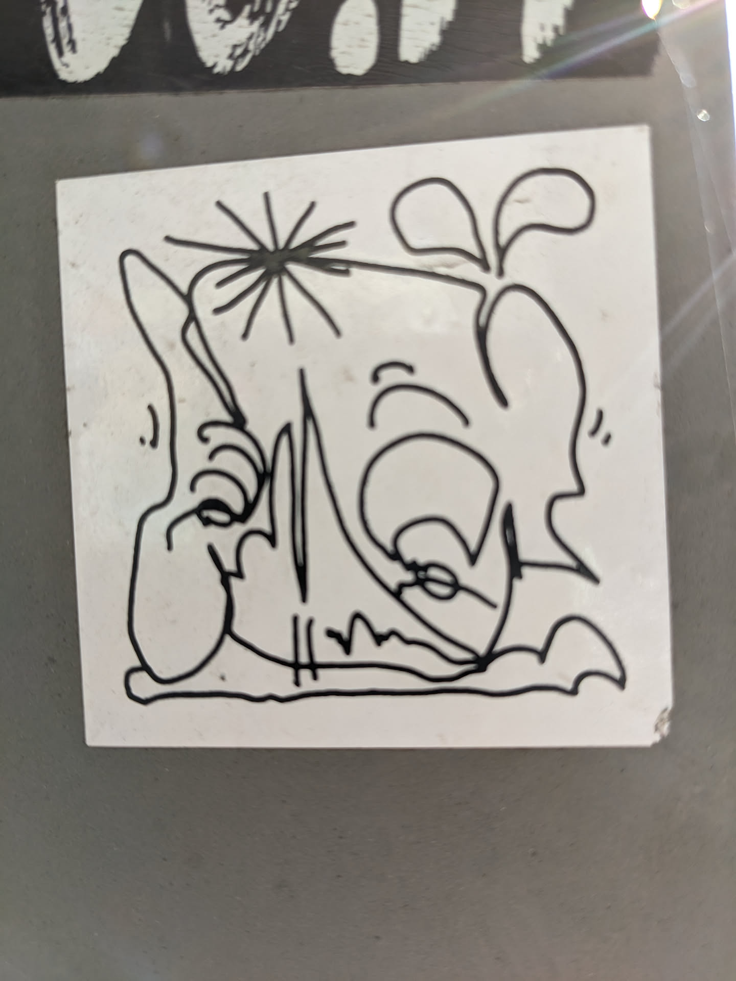 A white square sticker with a black and white line drawing of a cartoonish figure. The figure appears to be sitting or lounging, with a somewhat whimsical and abstract style. There's a small starburst element near the figure's head.