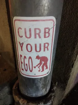 A sticker with a message to curb one's ego.  It features a person with a shovel, suggesting the act of cleaning or clearing something away. The sticker has a slightly worn appearance.