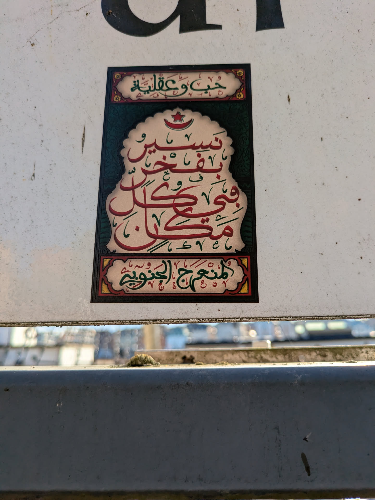 A rectangular sticker features intricate Arabic calligraphy. The text is in red, set against a decorative backdrop with ornate borders. The calligraphy is arranged in a visually appealing design with a crescent moon and star at the top. There are decorative elements and patterns around the central text, and a border that frames the entire design.