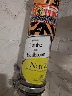 Street sticker A rectangular white sticker with a thin black border affixed to a vertical pipe. The center features black serif text that reads 'Stirbt die Laube stirbt Heilbronn'. The bottom portion of the sticker is partially covered by a circular yellow Baden-Württemberg promotional sticker.