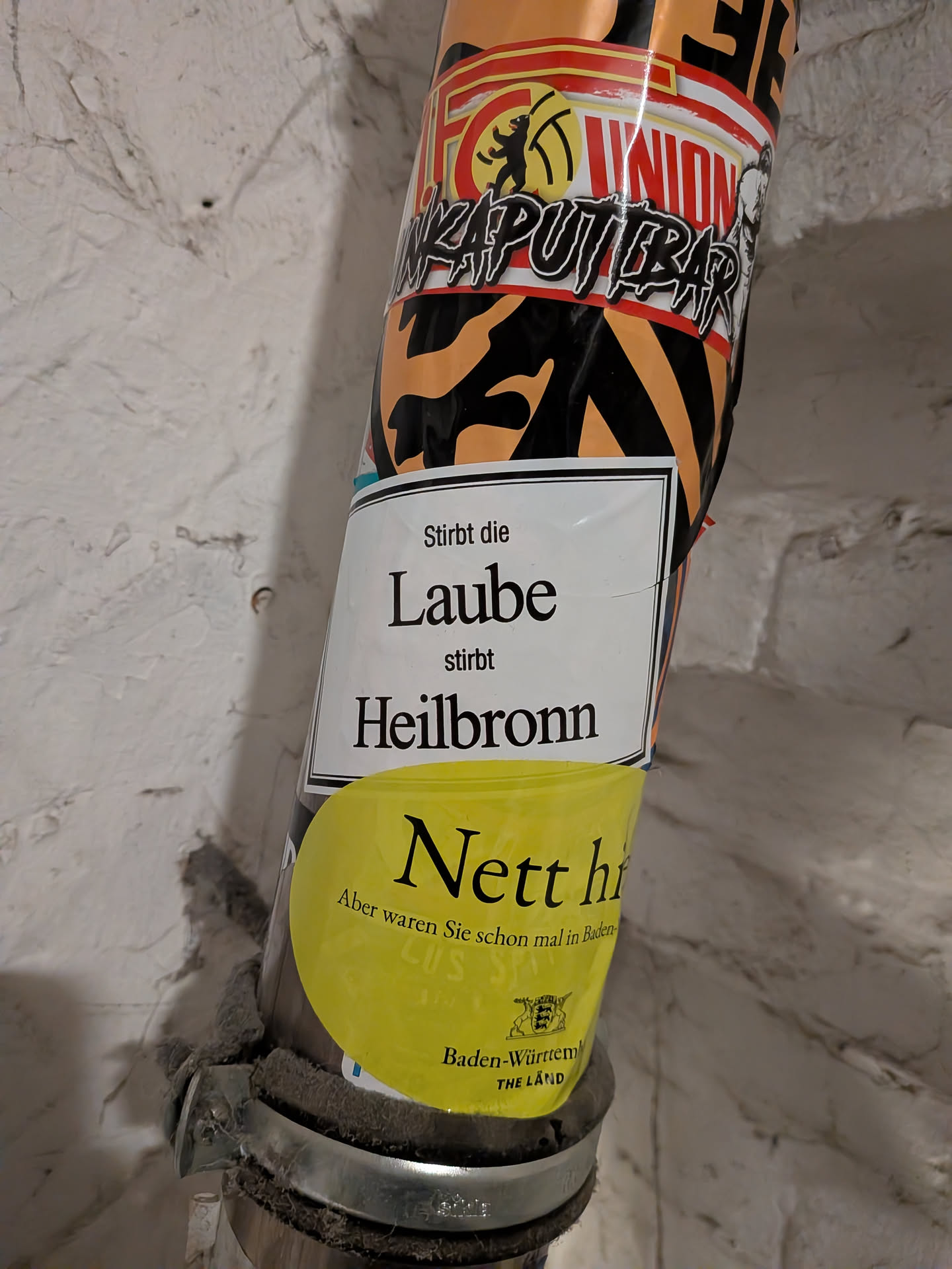 A rectangular white sticker with a thin black border affixed to a vertical pipe. The center features black serif text that reads 'Stirbt die Laube stirbt Heilbronn'. The bottom portion of the sticker is partially covered by a circular yellow Baden-Württemberg promotional sticker.
