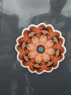 A round sticker featuring a detailed mandala design. The mandala is primarily in shades of brown and beige, with a small teal center. It has a complex, symmetrical pattern with a flower-like appearance. The sticker is affixed to a dark gray surface.