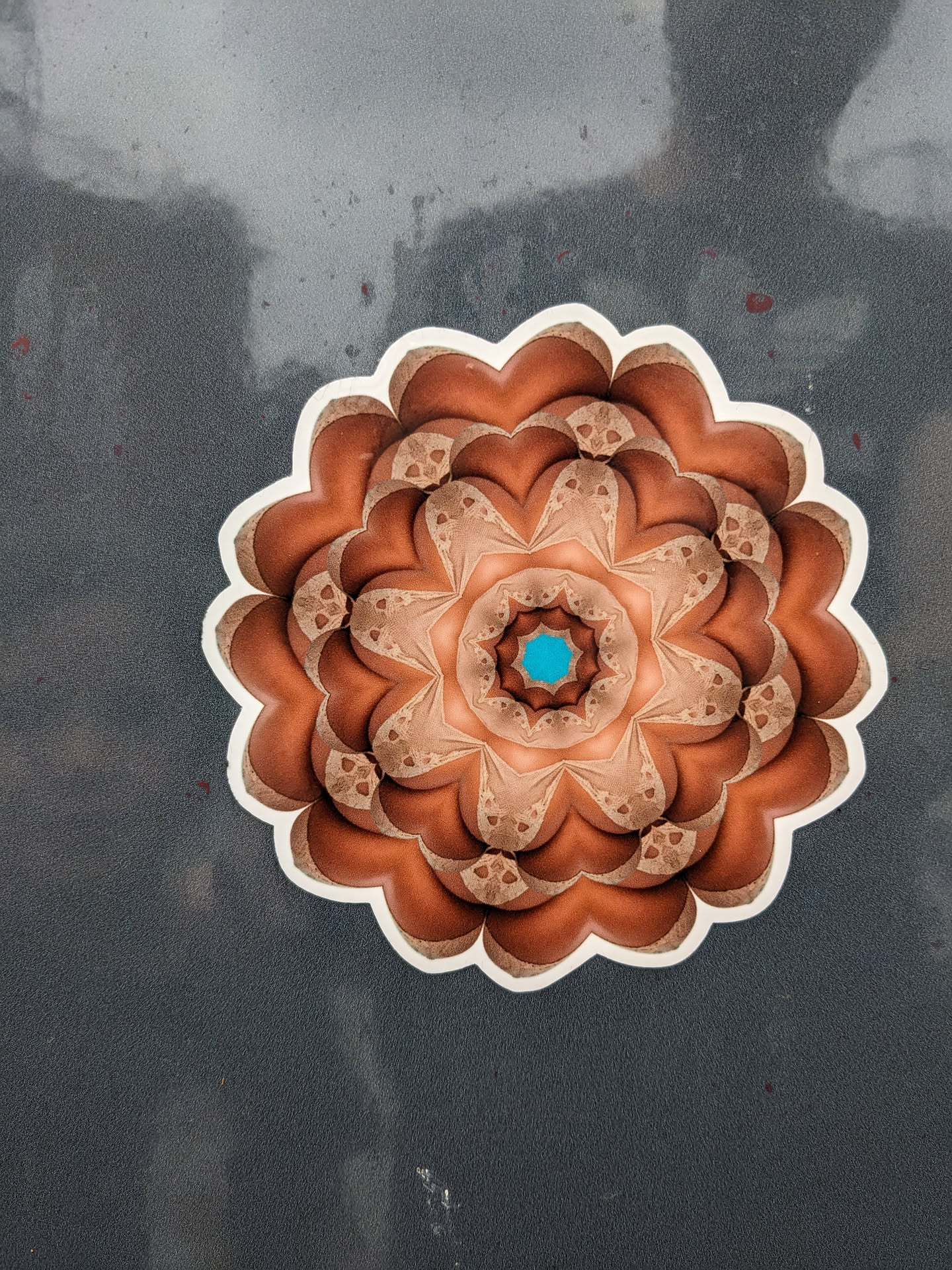 A round sticker featuring a detailed mandala design. The mandala is primarily in shades of brown and beige, with a small teal center. It has a complex, symmetrical pattern with a flower-like appearance. The sticker is affixed to a dark gray surface.