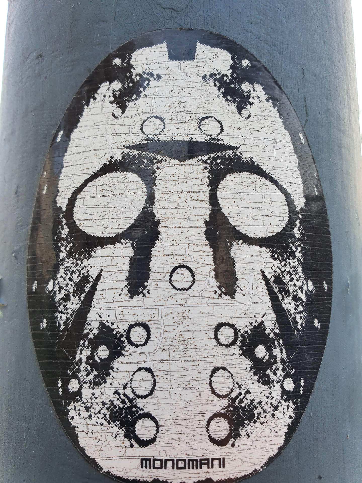 A black and white sticker featuring a stylized hockey mask, reminiscent of Jason Voorhees from Friday the 13th. The artwork has a gritty, almost pixelated texture, and is signed "Monomani" at the bottom. It's been placed on a grey metal pole.