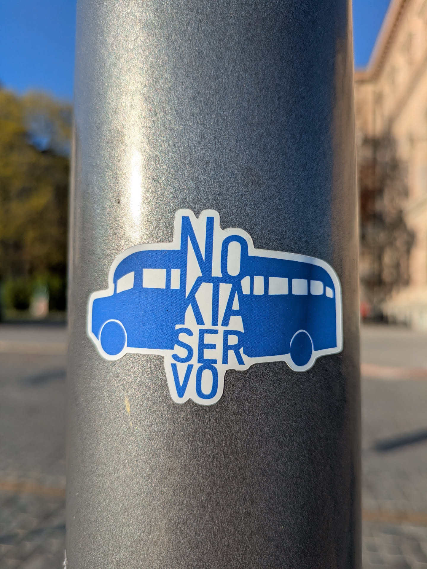 A blue and white sticker on a gray pole featuring a stylized bus with the text 'NO KIA SERVO' overlaid. The design is simple and somewhat humorous.