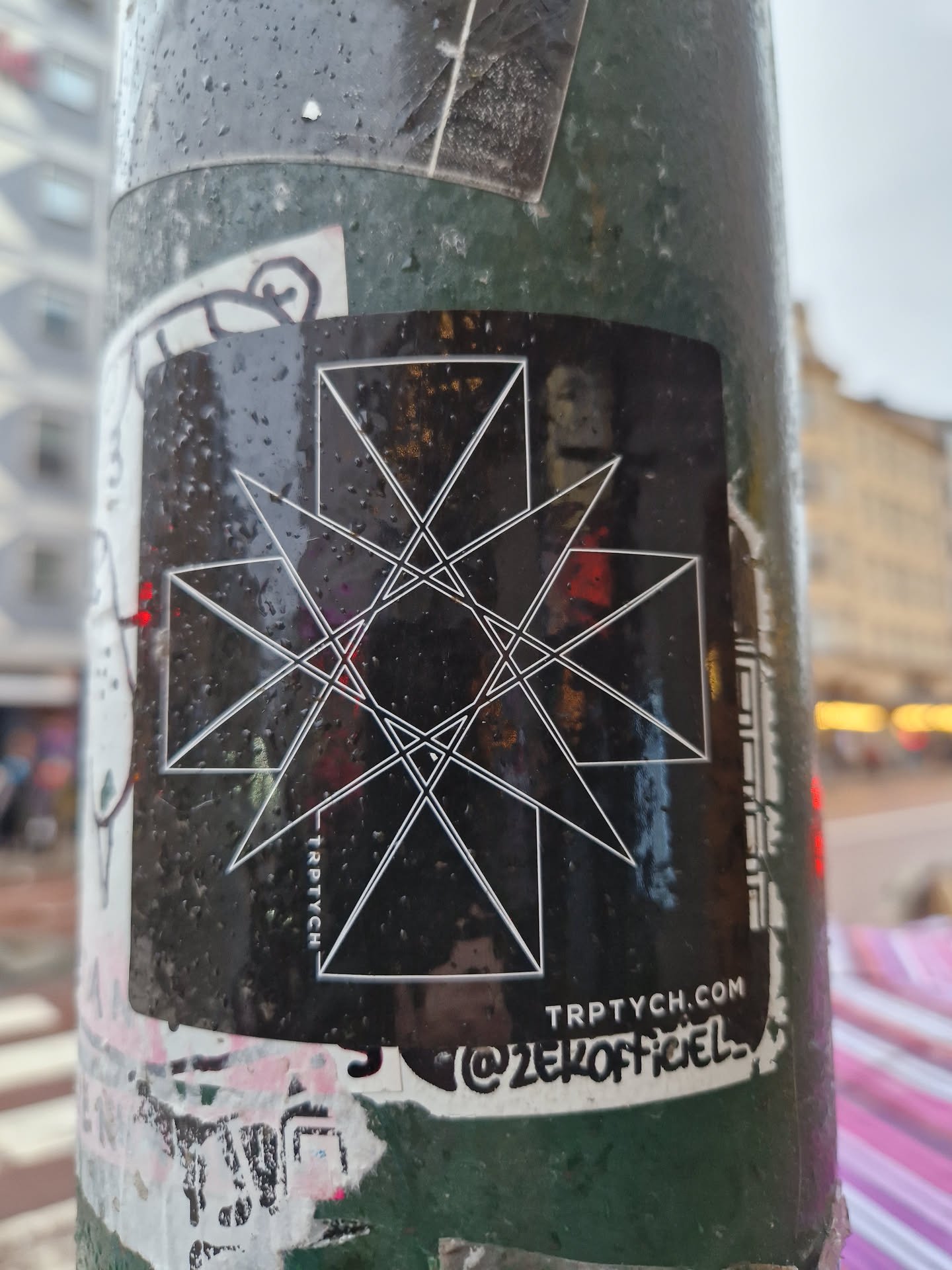 Black and white geometric design sticker with the website trptych.com and instagram handle @zer0officiel. The sticker is wet from rain and slightly reflective.