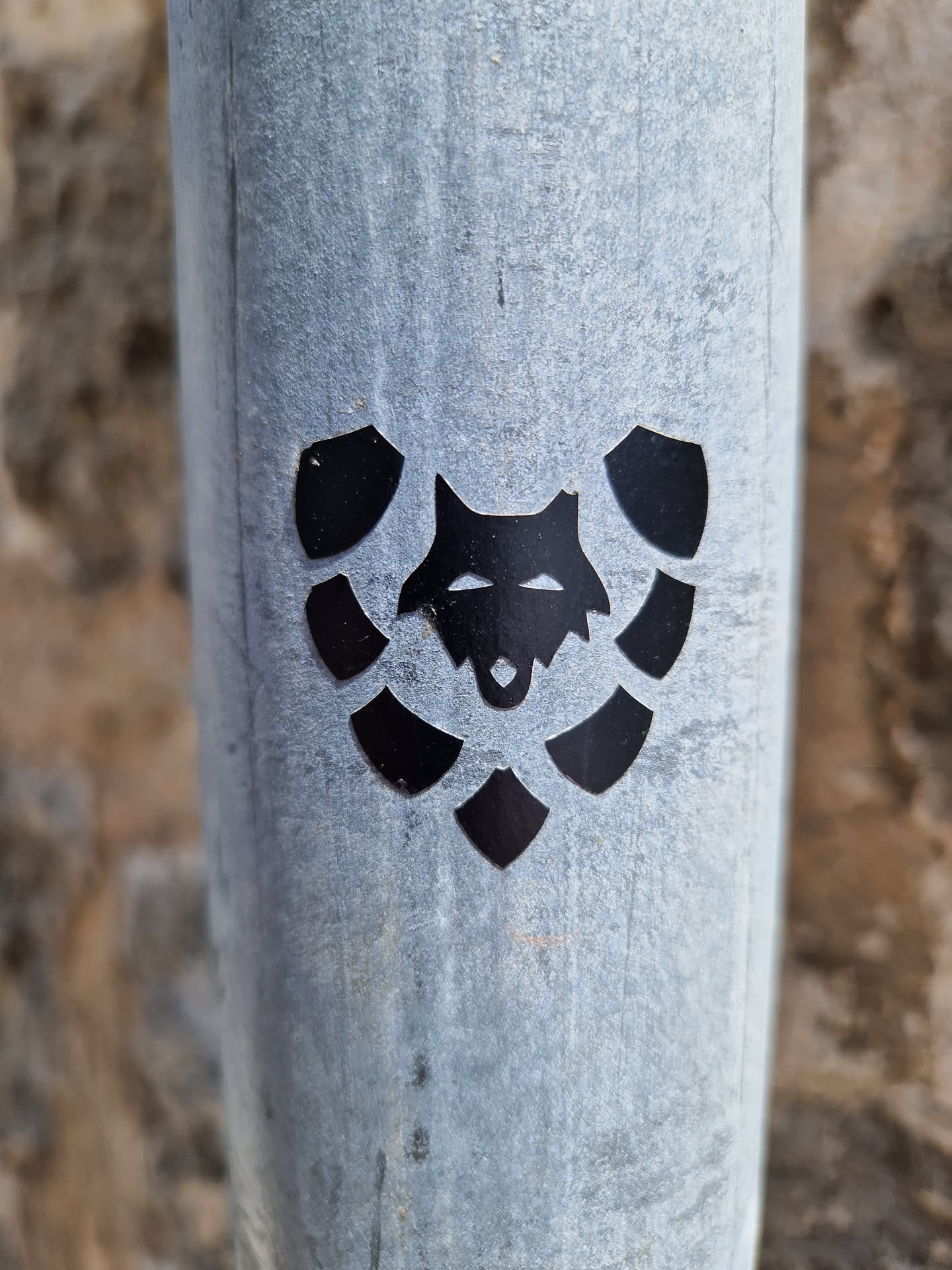 Close-up of a black vinyl sticker featuring a stylized wolf's head encircled by leaf-like shapes, adhered to a weathered gray metal pole. The sticker has a minimalist design with a strong graphic presence.