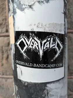 A black and white sticker is stuck to a pole. The sticker features the band name "OVERVALD" in an elaborate, thorny font. Below the band's name is the text "OVERVALD.BANDCAMP.COM".