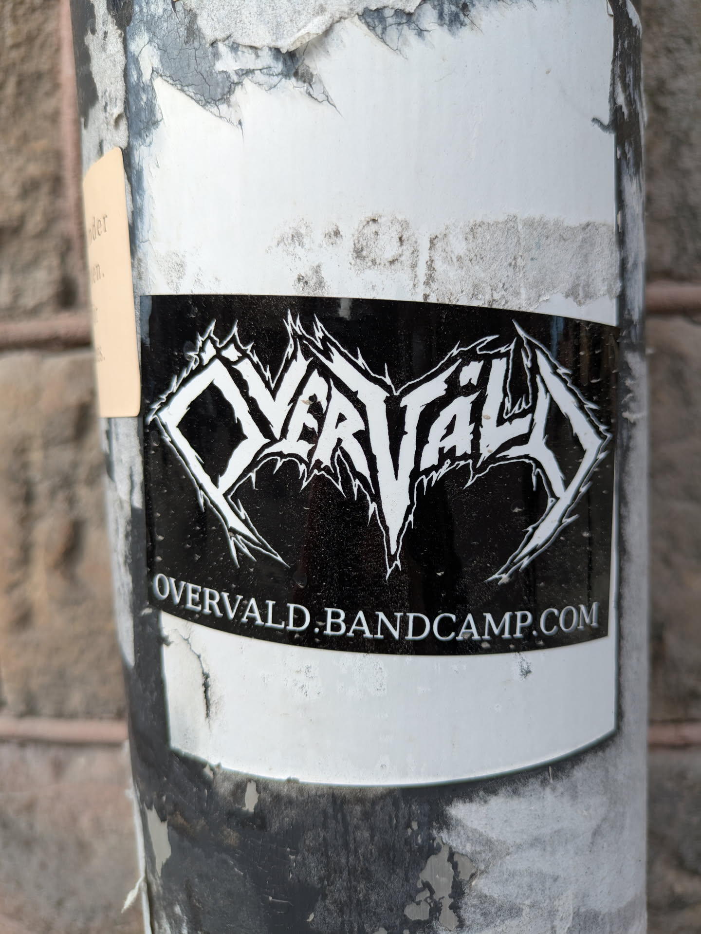 A black and white sticker is stuck to a pole. The sticker features the band name "OVERVALD" in an elaborate, thorny font. Below the band's name is the text "OVERVALD.BANDCAMP.COM".