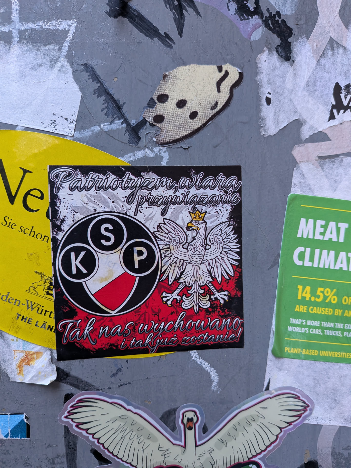 A square sticker featuring a stylized white Polish eagle with a golden crown, beak, and talons, set against a distressed background that transitions from white to red and black. To the left of the eagle, there is a circular emblem divided diagonally into white and red sections, containing three smaller black circles with white letters 'K', 'S', and 'P'. Above the eagle, white cursive text reads 'Patriotyzm, wiara, przywiązanie'. Below the eagle and emblem, more white cursive text states 'Tak nas wychowano i tak już zostanie!'. The overall design is strong and nationalistic, with a slightly gritty aesthetic.