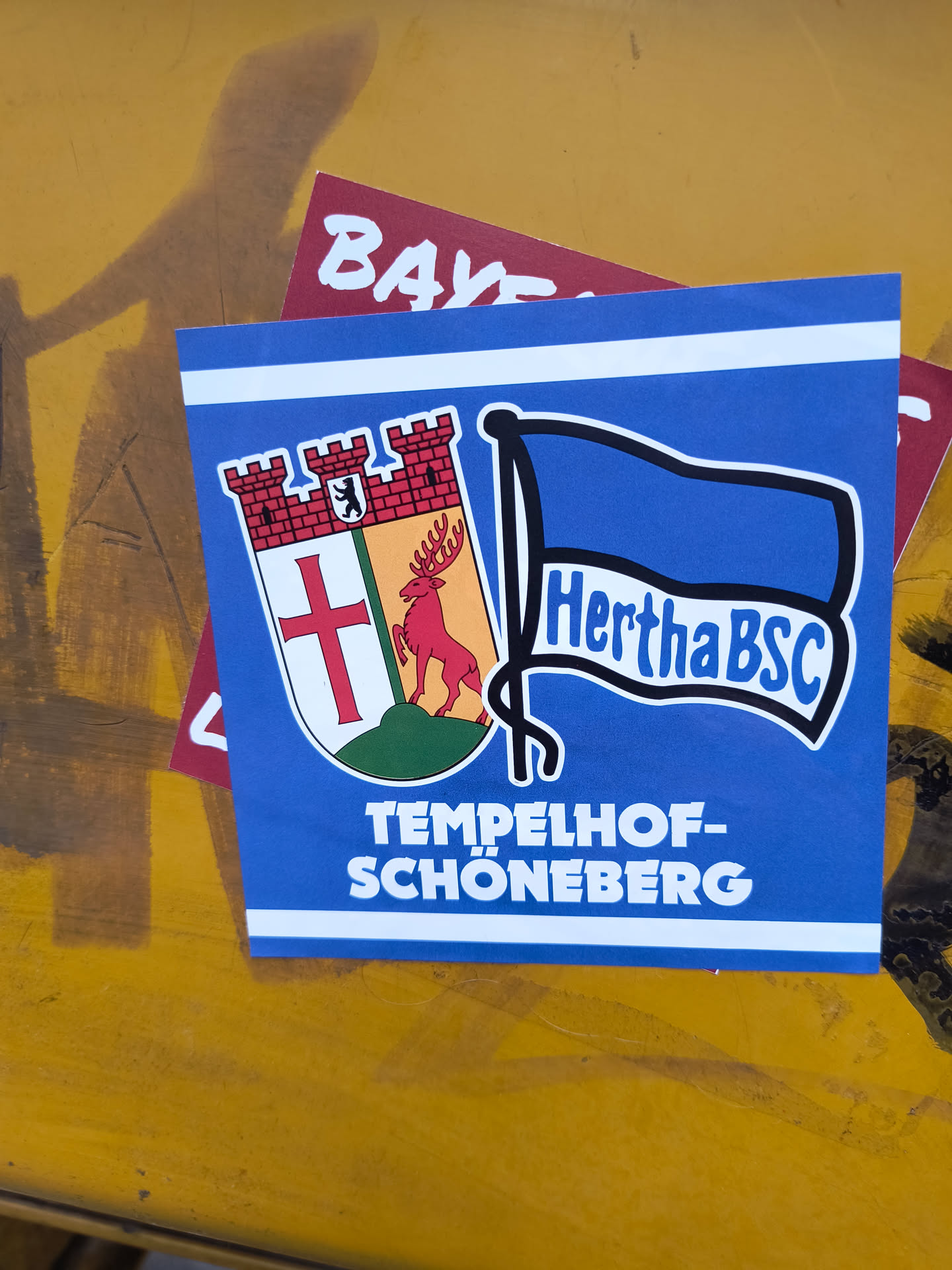 The sticker is predominantly blue with white horizontal stripes at the top. On the left side, it features a shield with a red cross on a white background, a red deer with antlers on an orange background, and a red brick castle with a bear emblem at the top. On the right side, a blue flag with the text 'Hertha BSC' in white is depicted. Below these elements, the text 'TEMPELHOF-SCHÖNEBERG' is written in large white capital letters.
