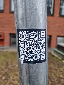 A black and white QR code sticker is affixed to a weathered metal pole. The pole appears to be outdoors, with blurred vegetation and a brick building visible in the background.