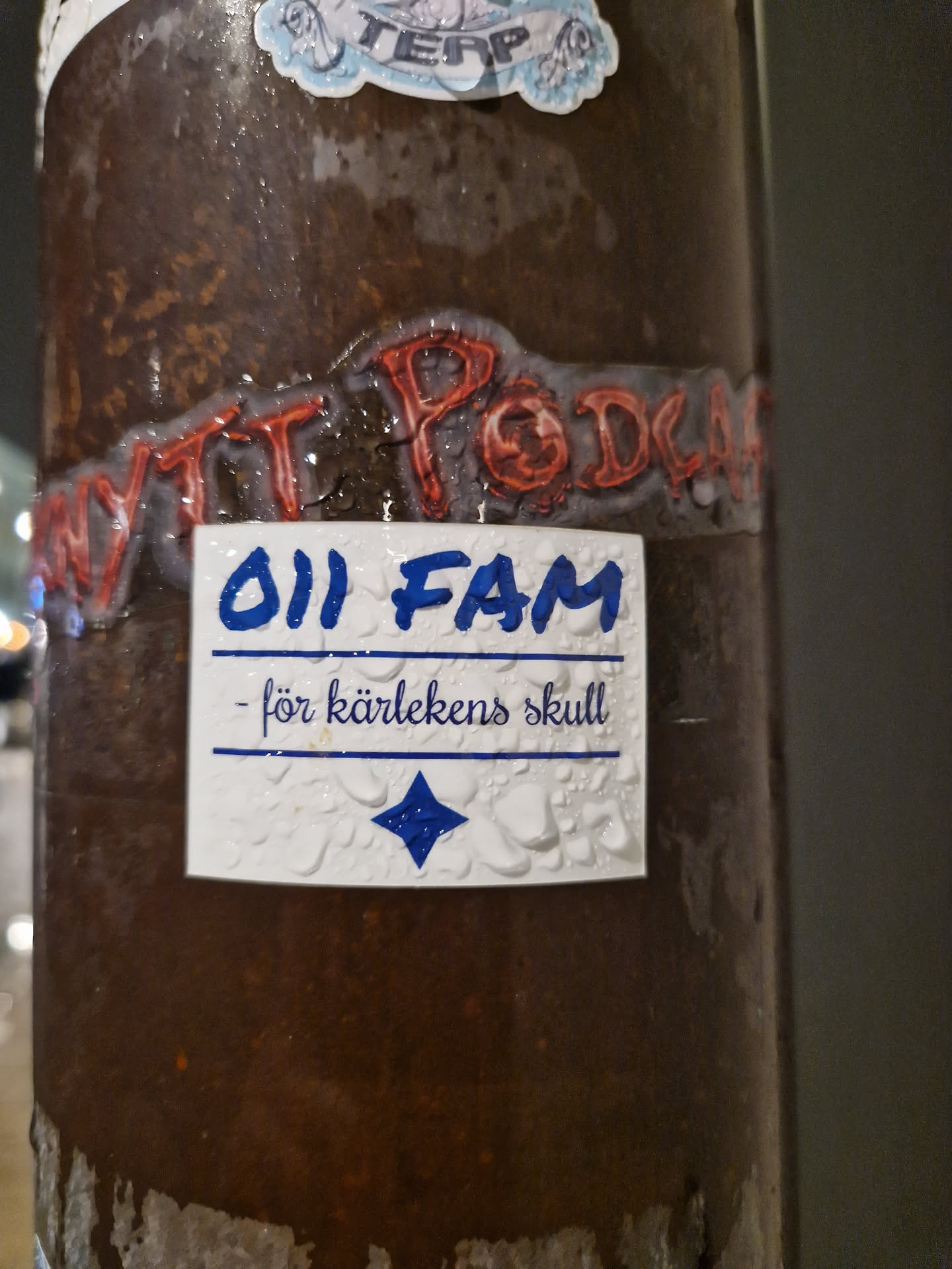 A small, white sticker with blue text and a blue diamond. The text says "011 FAM" and "- för kärlekens skull" (for the sake of love). The sticker is attached to a brown pole and has water droplets on it.