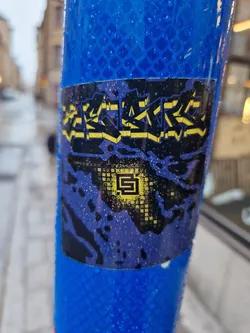 A dark purple and yellow sticker with graffiti-style text at the top and a pixelated design at the bottom featuring a stylized letter 'S' in a gold square. The sticker is wet, likely from rain. It's adhered to a blue reflective post.