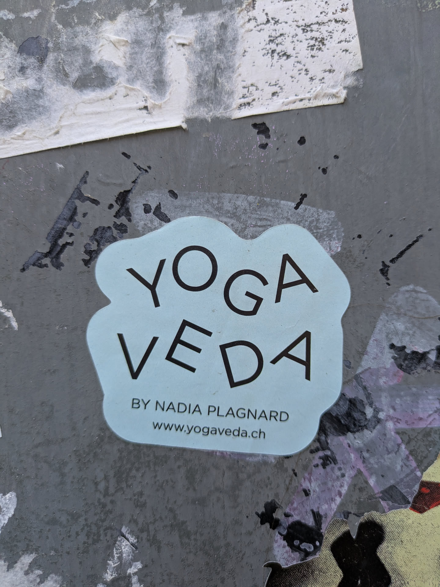 This sticker promotes Yoga Veda by Nadia Plagnard, with a website address provided. The sticker is light blue with dark text, placed on a weathered gray wall with remnants of other stickers.