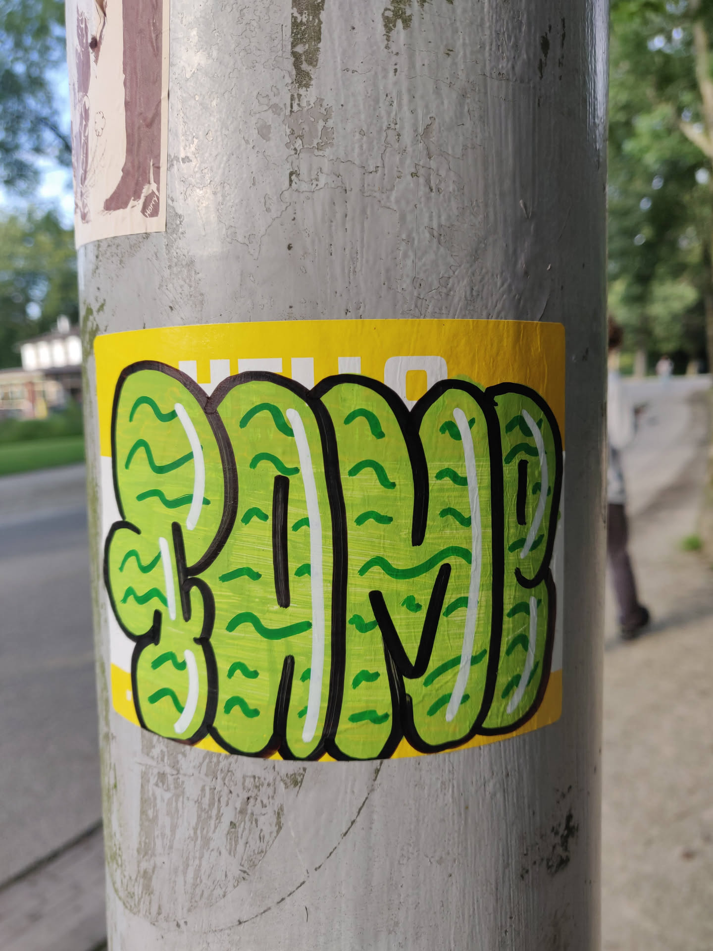 A green and yellow sticker with the word "FAM" written in a stylized, wavy font.  The letters are outlined in white and filled with a repeating wave pattern. The sticker is affixed to a grey pole.