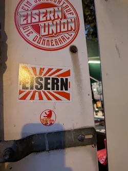 Street sticker A rectangular sticker positioned in the center of a painted metal pillar. The background design resembles a red and white rising sun motif with rays radiating from the center. Centered horizontally is the word 'EISERN' in bold, heavy, black capital letters outlined slightly in white. A light reflection is visible on the left half of the glossy sticker surface.