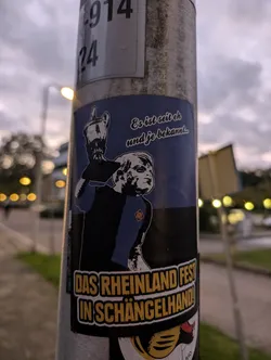 A sticker featuring a stylized illustration of a person holding a trophy, with text in German. The text at the top translates to: "It's been known since..." and the main text at the bottom says: "Das Rheinland Fest in Schängelhand!" which translates to "The Rhineland Festival in Schängelhand!"