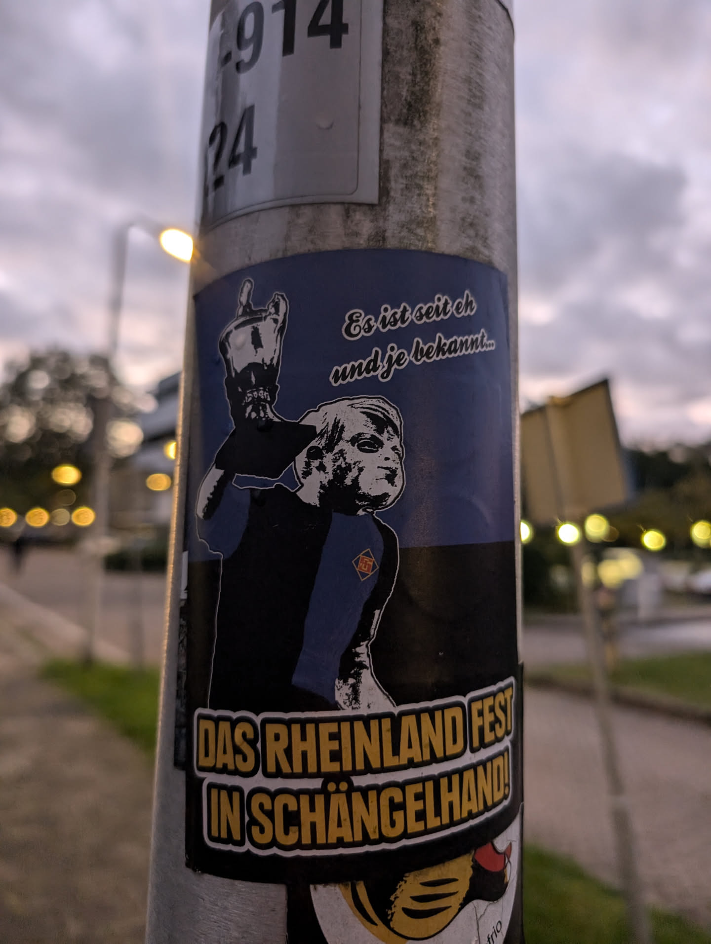A sticker featuring a stylized illustration of a person holding a trophy, with text in German. The text at the top translates to: "It's been known since..." and the main text at the bottom says: "Das Rheinland Fest in Schängelhand!" which translates to "The Rhineland Festival in Schängelhand!"
