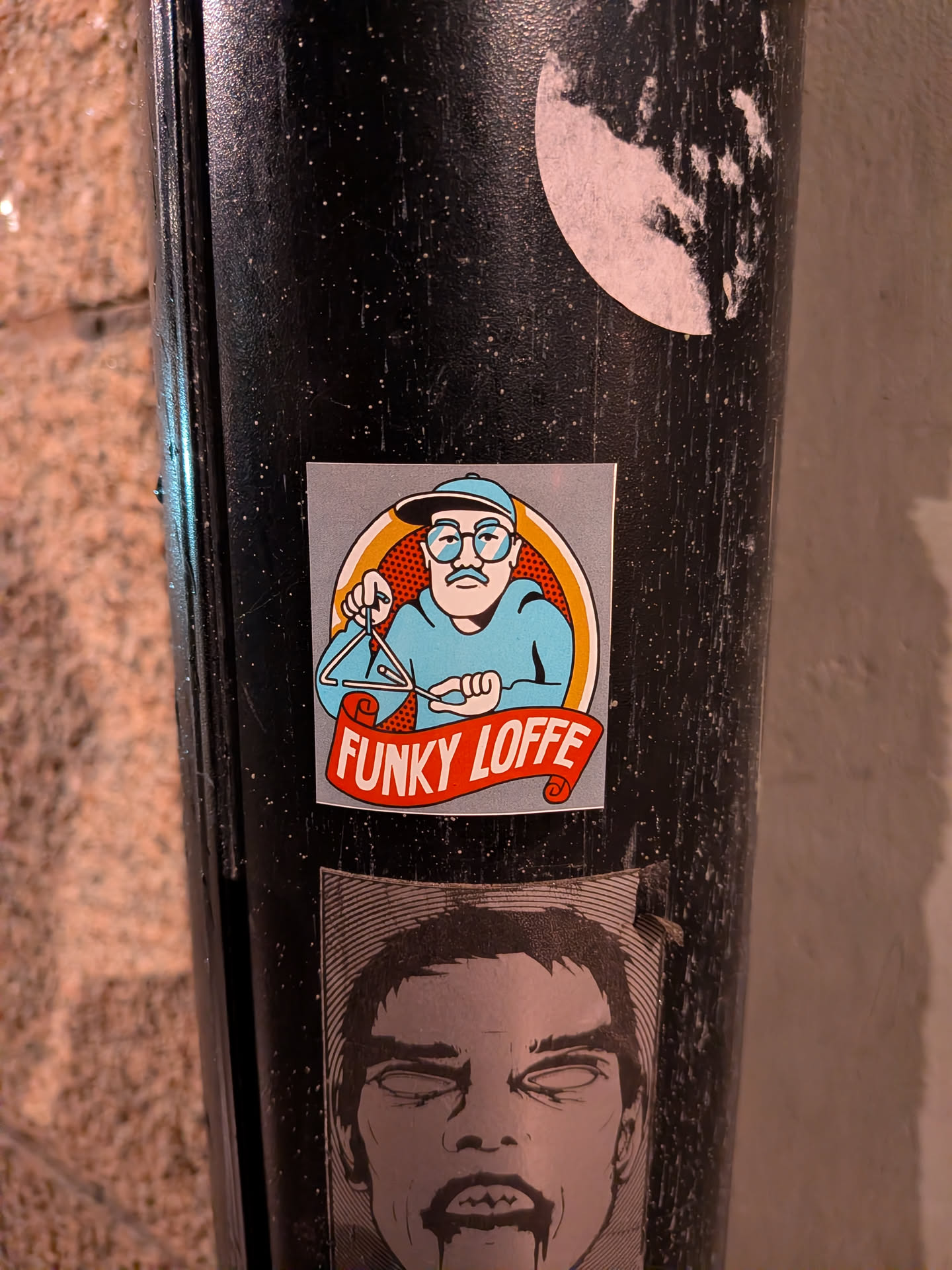 The sticker features a cartoon illustration of a man wearing a blue baseball cap and glasses, with a prominent mustache. He is dressed in a light blue shirt and is holding a triangle musical instrument with a beater. A red banner with white text wraps around the bottom of the illustration. The background of the sticker is a light gray with a circular orange and yellow gradient behind the man's head, and a red dotted pattern within the orange circle. The overall style is reminiscent of retro comic art.