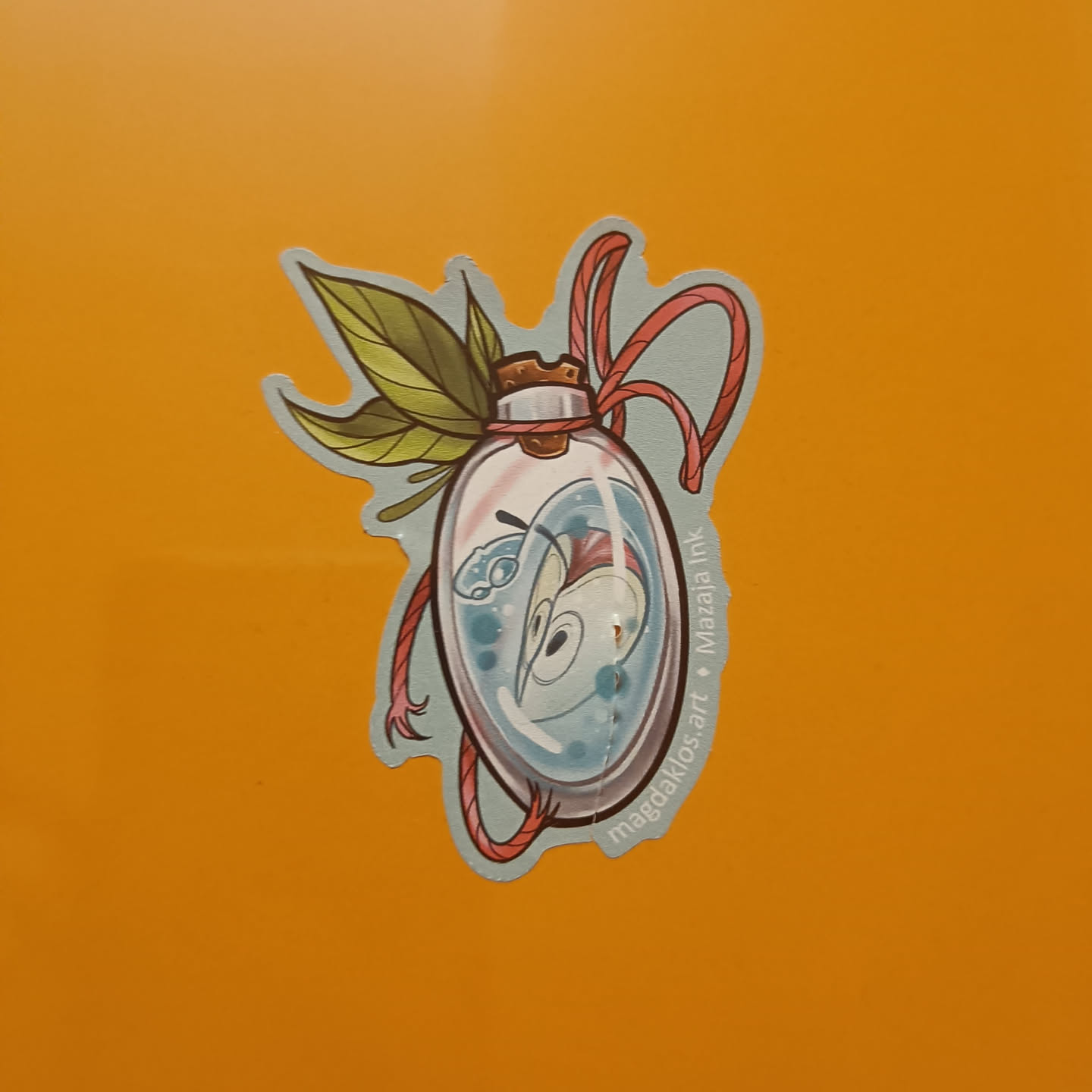This delightful sticker showcases a unique illustration of a creature sealed within a glass bottle, adorned with leaves and a red string.  The style is whimsical and charming, with soft pastel colors and delicate linework. The artist's name and social media handle are subtly integrated into the design.