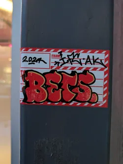 A red and yellow sticker with graffiti-style lettering spelling out the word 'BETS'. It's designed to look like a shipping label with '2024' and a tag written above the word 'BETS'. The tag on the top appears to say 'FROM  ...AK'.