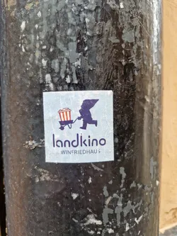 A sticker featuring a minimalist design of a figure pushing a popcorn cart, with the text 'landkino WINFRIEDHAUS' underneath. The sticker is affixed to a weathered, dark-colored pole.