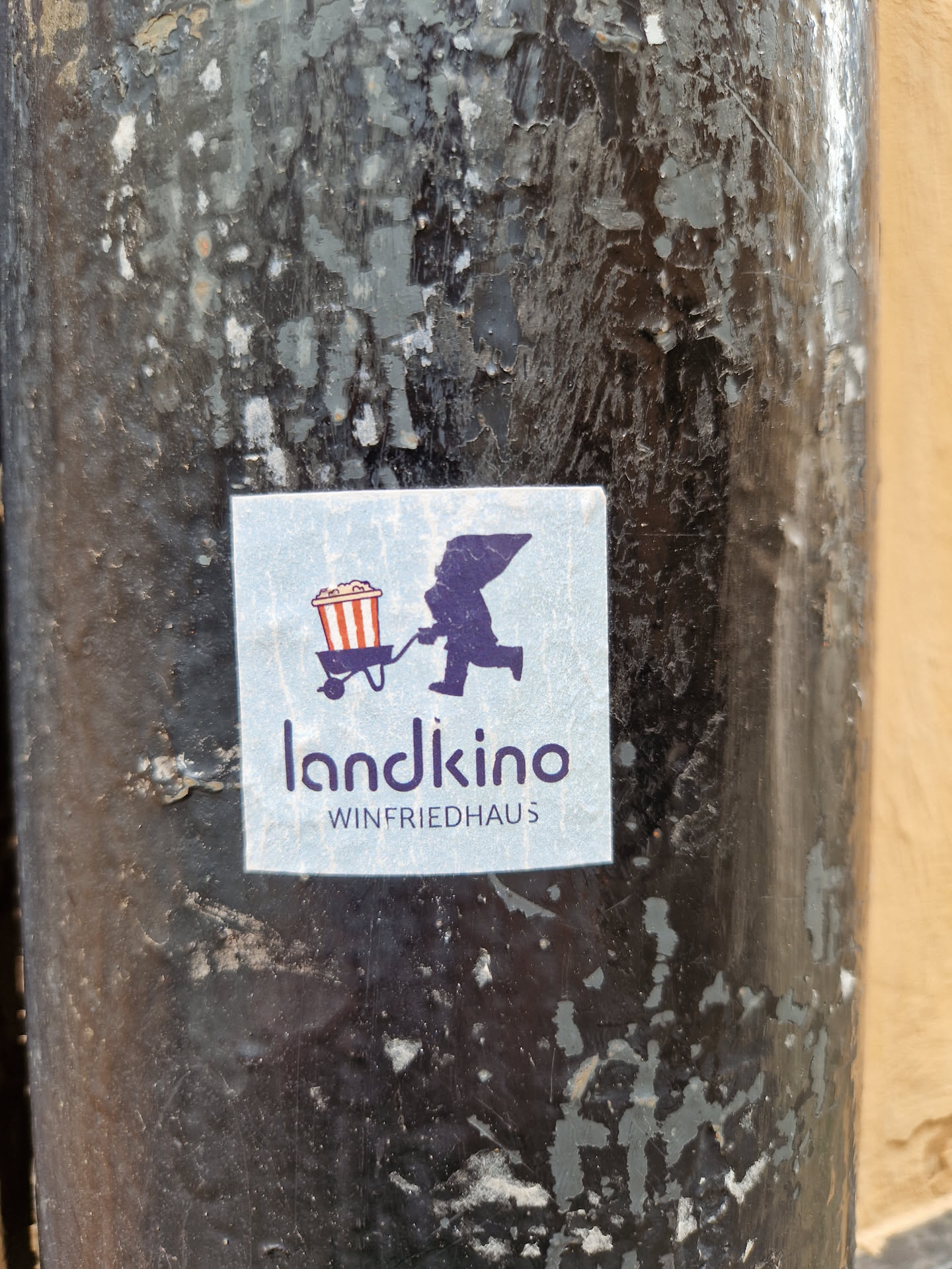 A sticker featuring a minimalist design of a figure pushing a popcorn cart, with the text 'landkino WINFRIEDHAUS' underneath. The sticker is affixed to a weathered, dark-colored pole.