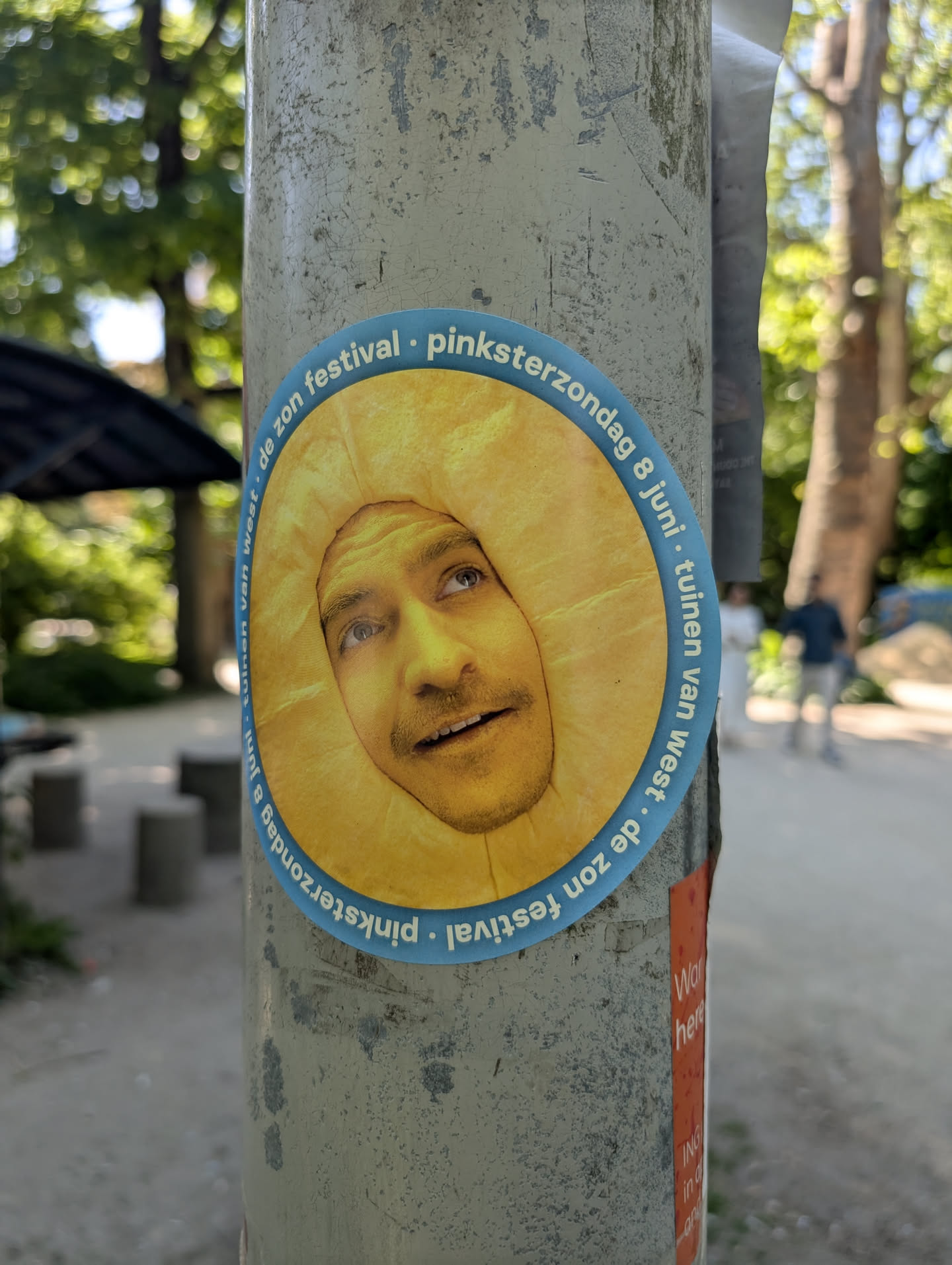 A round sticker on a pole shows a man's face framed by a bright yellow sun shape. The sticker advertises a festival, with text circling the edge. The background is a blurry outdoor scene with trees and a path.