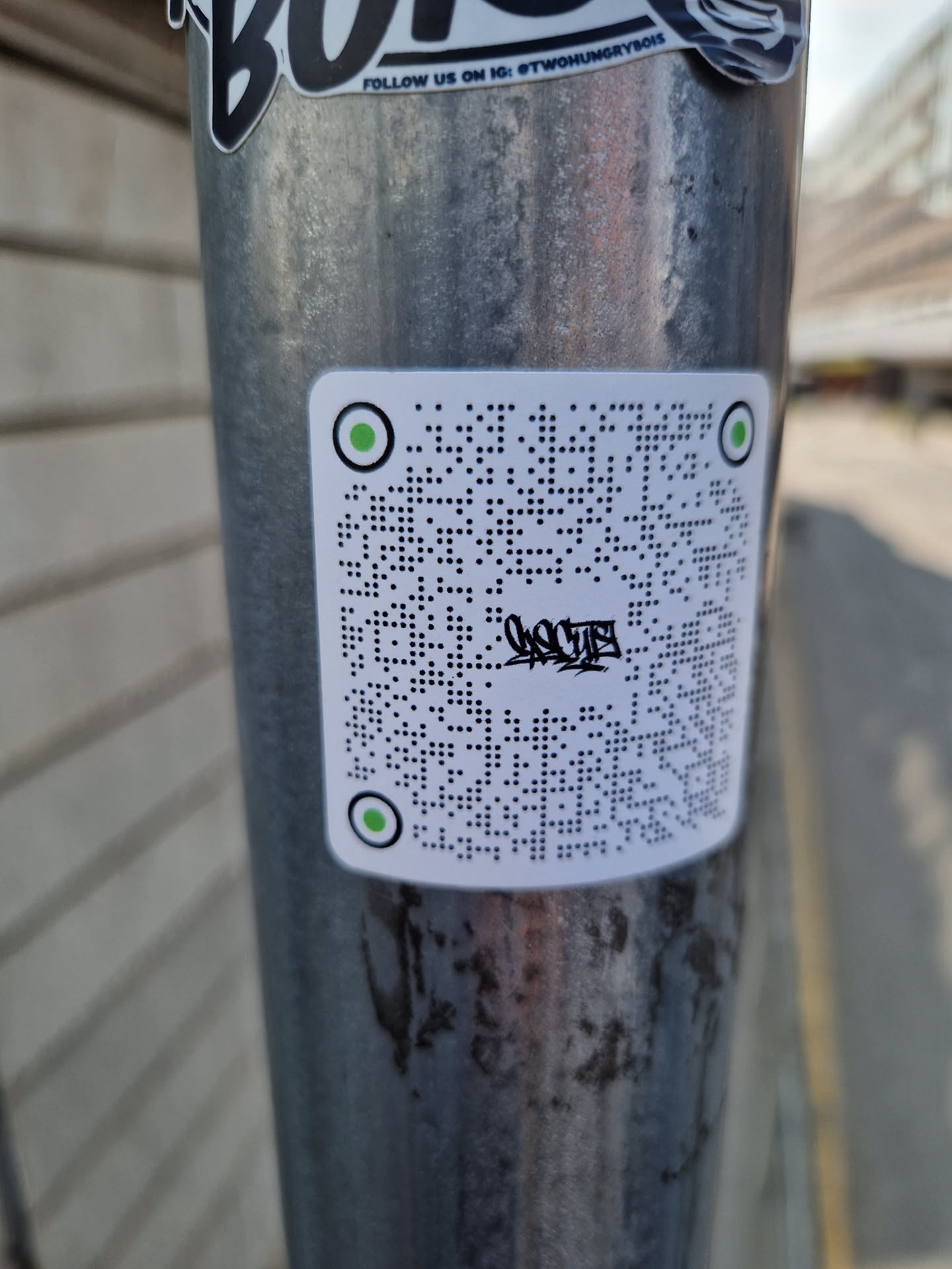 A white square sticker with a QR code in the center and green circles on each corner. There is some black paint on the QR code.  The sticker is affixed to a metal pole.