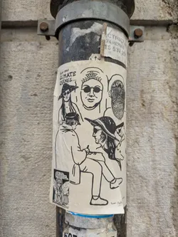 The sticker features a collage of black and white drawings on a neutral background. The drawings include people wearing hats and glasses, and a steampunk-esque mask. The text reads 'Climate Change...' and includes the signature 'Juan Salc'. There's another smaller sticker on the lower left, depicting a fairy-like character. The sticker is affixed to a gray, industrial metal pipe.