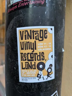 A sticker advertising Vintage Vinyl Records in Lund, Sweden.  The sticker features illustrations of two figures carrying vinyl records and indicates that they buy and sell LPs and records. Contact information is included.